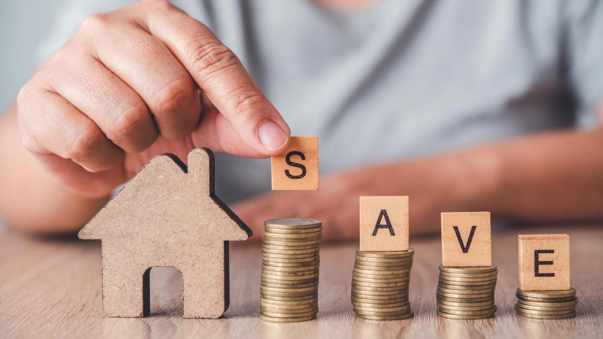 Hand placing a wooden block with "S" on a stack of coins; other blocks spell "SAVE" on coin stacks. House-shaped figure on the left.