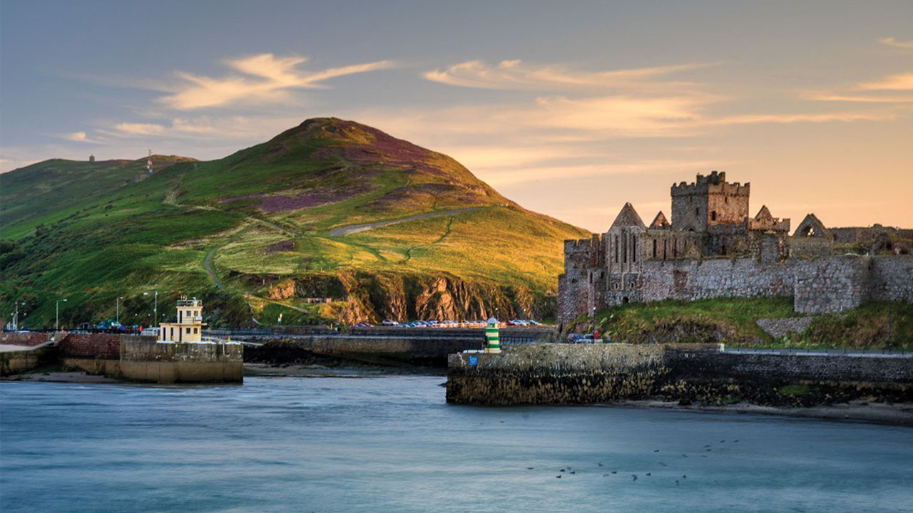 Coastal scene with a historic stone ruin near the shore, a small harbor with a lighthouse, and a green hill in the background under a partly cloudy sky.