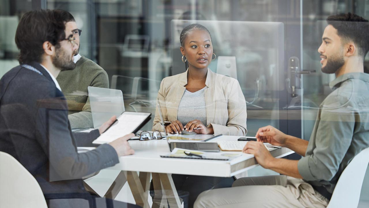 Four people sit around a table in an office, engaged in discussion. One person holds a tablet, while papers and a laptop are on the table. The meeting room has glass walls.