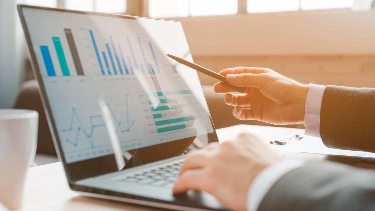 Person pointing at charts on a laptop screen with a pen, analyzing data in a bright office setting.