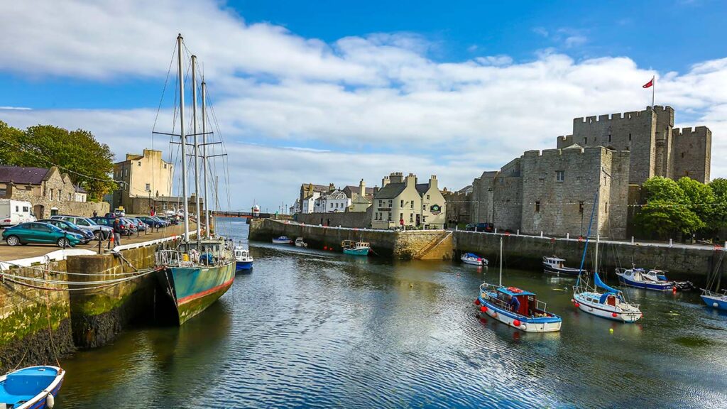 A small harbor with several boats, a stone castle, and surrounding buildings under a partly cloudy sky.