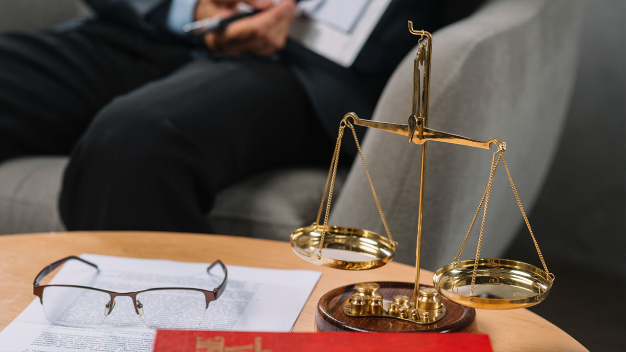 A brass balance scale on a table with documents, a pair of glasses, and a red book. A person holding a pen and notebook is seated in the background.