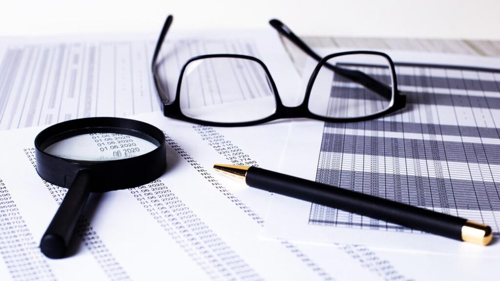 A pair of glasses, a magnifying glass, and a black pen rest on sheets of numerical data and graphs.