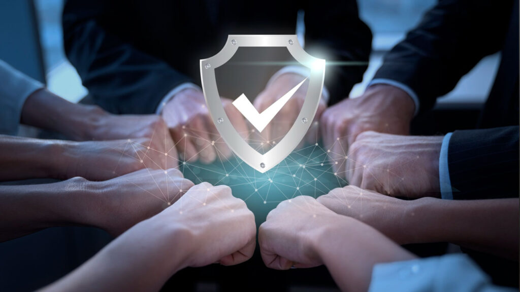 Several hands form a fist bump under a digital shield icon with a check mark, symbolizing teamwork and security.