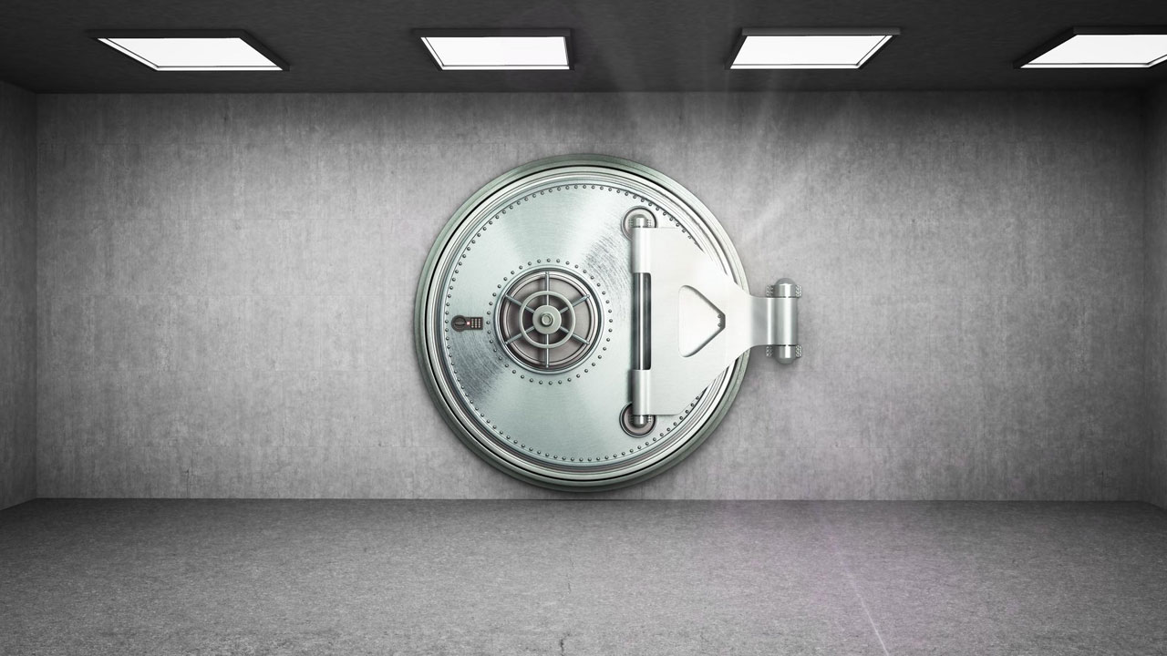 Large metal bank vault door in a concrete room with overhead lights, slightly ajar with light streaming in.